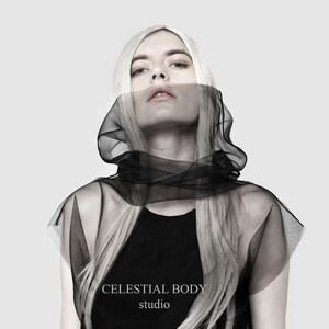 May include: A woman with long blonde hair wears a black sheer hooded top with the text "CELESTIAL BODY studio" on the bottom.