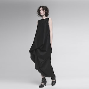 May include: A woman wearing a black sleeveless dress with a draped design. The dress has a loose, flowing silhouette and falls to her ankles. She is wearing black heeled shoes.