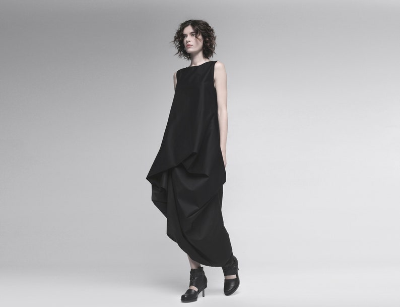 May include: A woman wearing a black sleeveless dress with a draped design. The dress has a loose, flowing silhouette and falls to her ankles. She is wearing black heeled shoes.
