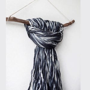 May include: A decorative wall hanging featuring a natural wooden branch with a bundle of dark blue, gray, and white yarn tied to it. The yarn hangs down in a fringe, creating a textured, minimalist design. The branch is suspended by a thin string.