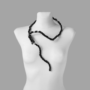 May include: A black necklace with rectangular beads is draped on a white mannequin. The necklace has a unique, flowing design, with a few silver-colored beads interspersed throughout. The background is a solid gray.