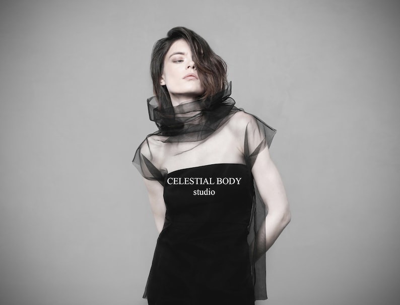 May include: A woman wearing a black strapless dress with a sheer black overlay. The overlay is gathered at the neck and falls to the shoulders. The text "CELESTIAL BODY studio" is printed on the dress.