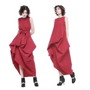 May include: Two women wearing red sleeveless dresses with a gathered, asymmetrical design. The dresses have a simple, round neckline and a fitted bodice. The women are wearing black heeled shoes.