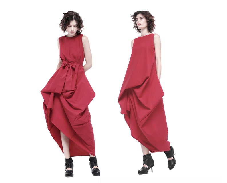 May include: Two women wearing red sleeveless dresses with a gathered, asymmetrical design. The dresses have a simple, round neckline and a fitted bodice. The women are wearing black heeled shoes.