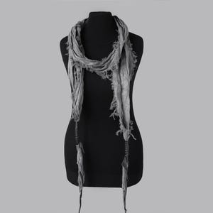 May include: A grey scarf with a distressed, fringed design is draped on a black mannequin. The scarf has long, dangling ends and a textured appearance, suggesting a lightweight fabric. The scarf is a fashion accessory.