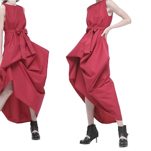 May include: Two women wearing red sleeveless dresses with a bow at the waist. The dresses have a gathered, asymmetrical design. The women are wearing black ankle boots.