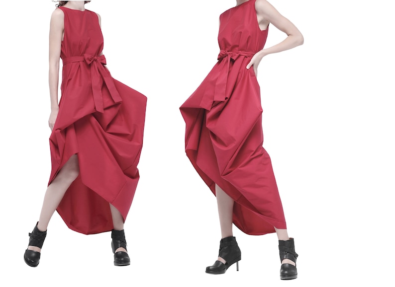 May include: Two women wearing red sleeveless dresses with a bow at the waist. The dresses have a gathered, asymmetrical design. The women are wearing black ankle boots.