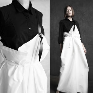 May include: A white, flowing, asymmetrical dress with a black ribbon detail. The dress is worn over a black button-down shirt.