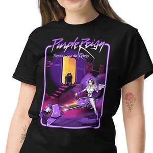 Puede incluir: Camiseta negra con un diseño gráfico que muestra el texto "Purple Reign". El diseño presenta una escena futurista con una figura en blanco y morado, enmarcada en morado. La camiseta la lleva una persona.