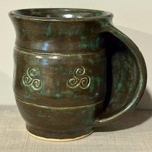 May include: A handmade ceramic mug with a dark brown glaze and green accents. The mug features a large handle and two embossed Celtic knot designs. The mug is approximately 5 inches tall.