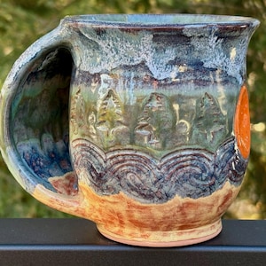 May include: A ceramic mug featuring a hand-thrown design with a large, integrated handle. The exterior displays a relief pattern of pine trees above wavy lines, finished in a glaze of blue, green, and tan tones with a circular orange accent.