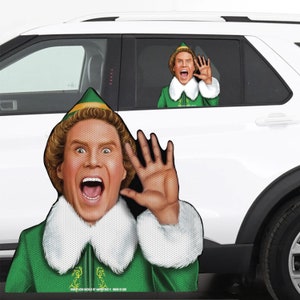 May include: A Buddy the Elf car window cling featuring Buddy's face and a green elf hat. Buddy is wearing a green suit with a white collar and is holding up his hand. The cling is on a white car.