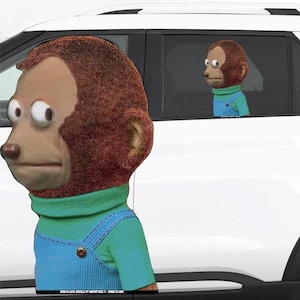 May include: A white car with a brown and green monkey puppet with large eyes looking out the window. The monkey is wearing a blue overall and a green shirt.