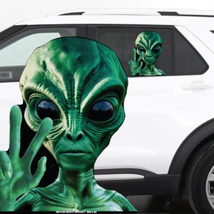 May include: Green alien decal with large black eyes and a peace sign hand gesture. A second alien decal is visible in the car window. The decals are placed on a white vehicle.