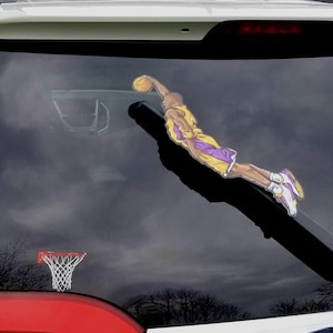 May include: A car window decal featuring a basketball player in a yellow and purple uniform, mid-dunk. The decal is positioned above a small basketball hoop. The background is a cloudy sky.