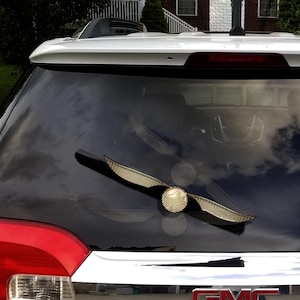 May include: A golden Snitch car decal, a popular item from the Harry Potter series, is affixed to the rear window of a vehicle. The decal features a round ball with wings, reflecting the sky and surrounding environment.