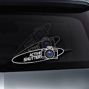May include: A black car with a white sticker on the back window. The sticker features a camera with a blue lens and the text "ACTIVE SHUTTER".