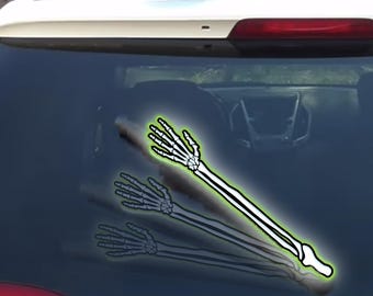 May include: A white and black skeleton arm car decal with a green outline is applied to a car window. The decal is a humorous Halloween decoration. The arm is angled diagonally across the window.