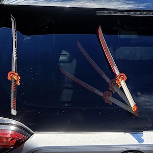 May include: A set of decorative katana-style car window decals. The decals feature a red and black color scheme with intricate details, including a white blade and orange hilt. The decals are designed to be applied to a car's rear window.