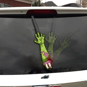 May include: A green zombie arm sticker with exposed bone and flesh, blood, and a torn sleeve. The sticker is applied to the rear window of a vehicle. The image shows a zombie theme.
