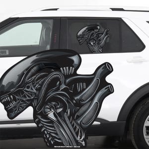 May include: Black and gray Alien face decal, with detailed shading and a textured appearance. The decal is applied to the window of a white vehicle, with a smaller version on the rear window. The design features the iconic creature from the Alien film series.