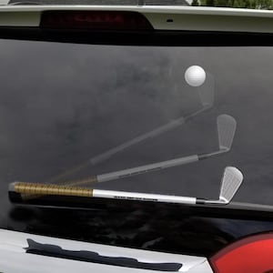 May include: A golf-themed car window decal featuring a golf ball in mid-air and a golf club. The club has a gold and black grip and a silver head. The decal is designed to look like a golf swing.