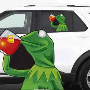 May include: A green frog character with a large, wide-eyed expression is drinking from a glass of red liquid. The frog is wearing a green, textured shirt. The image is a sticker that can be applied to a car window. The sticker is partially visible on the car window behind the frog. The sticker on the car window shows the same frog character drinking from a glass of red liquid.