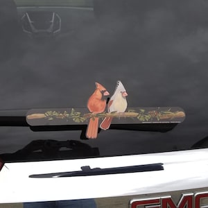 May include: A decorative car window sticker featuring two cardinal birds perched on a branch with leaves and berries. One cardinal is orange and the other is white and gray. The sticker is designed to be placed on a car's rear window.