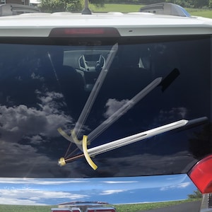 May include: A car window with a decal of a sword. The sword has a silver blade, a gold crossguard, and a brown handle. The background shows a cloudy sky and the interior of the car.