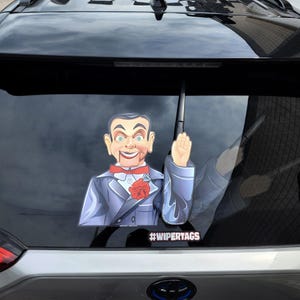 May include: A car window decal featuring a ventriloquist dummy with blue eyes, wearing a blue suit, red bow tie, and a red rose. The decal includes the text #WIPERTAGS.