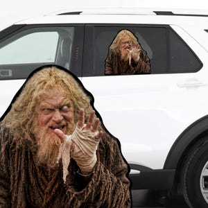 May include: A car window decal featuring a character with long, messy blonde hair and a beard, wearing a textured brown garment. The character has one eye looking upward and is reaching forward with a hand wrapped in light-colored, frayed bandages.