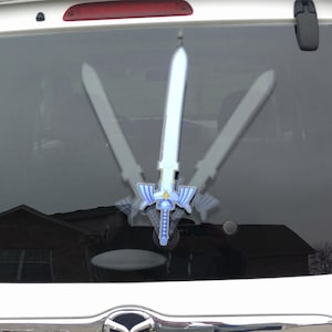 May include: A car window decal featuring a stylized sword design with blue and white accents. The decal is mounted on the rear window of a vehicle, with two mirrored sword images on either side of the central sword.