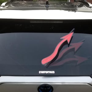 May include: A red and black arrow-shaped decal is affixed to the rear window of a vehicle. The decal has a curved design with a shadow effect, and the text "#WIPERTAGS" is below it.