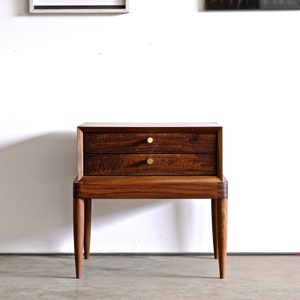 May include: A dark wood nightstand with two drawers and brass pulls. The nightstand has a simple, modern design and is sitting on a concrete floor.