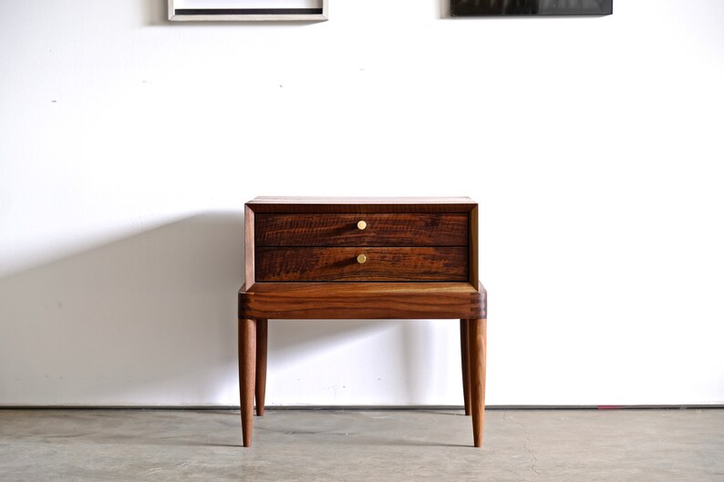 May include: A dark wood nightstand with two drawers and brass pulls. The nightstand has a simple, modern design and is sitting on a concrete floor.