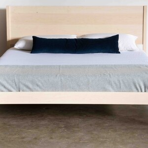 K&ouml;nnte beinhalten: Ein einfacher Holzbettgestell mit einer hellen Holzoptik. Das Bett ist mit wei&szlig;en Laken und einem blauen Kissen bezogen.