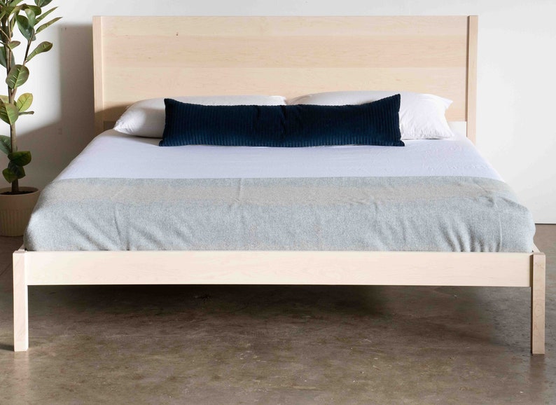 K&ouml;nnte beinhalten: Ein einfacher Holzbettgestell mit einer hellen Holzoptik. Das Bett ist mit wei&szlig;en Laken und einem blauen Kissen bezogen.