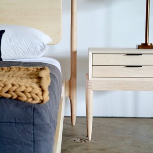 May include: A light wood nightstand with two drawers and four legs. The nightstand is next to a bed with a white pillow and a brown knitted throw blanket.