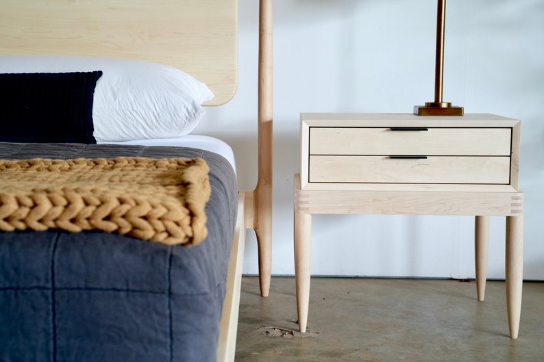 May include: A light wood nightstand with two drawers and four legs. The nightstand is next to a bed with a white pillow and a brown knitted throw blanket.