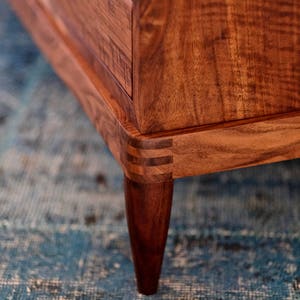 May include: Close-up of a wooden dresser with a dark brown finish. The dresser has a simple design with a few drawers and tapered legs. The dresser is sitting on a blue rug.