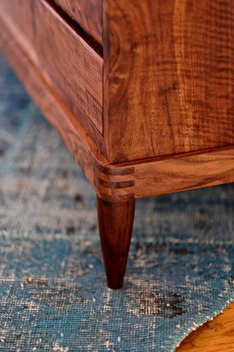 May include: Close-up of a wooden dresser with a dark brown finish. The dresser has a simple design with a few drawers and tapered legs. The dresser is sitting on a blue rug.