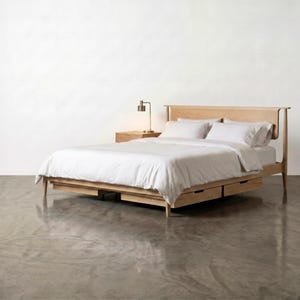 May include: A light-colored wooden bed frame with a headboard, two drawers, and a nightstand. The bed is covered with white bedding and pillows. A small lamp sits on the nightstand.