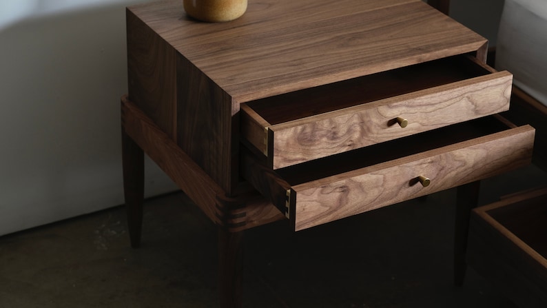 May include: A wooden nightstand with two drawers and brass knobs. The nightstand is made of walnut wood and has a dark brown finish.