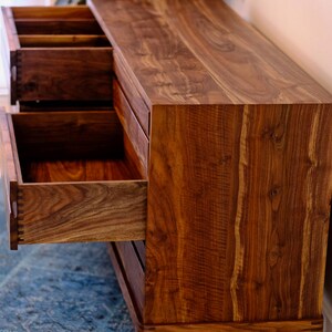 May include: A wooden dresser with four drawers, made of dark walnut wood. The dresser has a simple design with straight lines and a slightly tapered base. The drawers are open, revealing the wood grain inside.