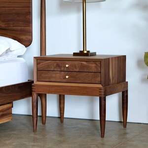 May include: A wooden nightstand with two drawers and four legs. The nightstand is made of dark wood and has brass drawer pulls. The nightstand is next to a bed with white bedding.