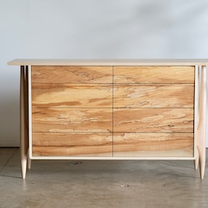 Op de afbeelding: Een lichte houten commode met zes laden en taps toelopende poten. De commode heeft een natuurlijke houten afwerking en een eenvoudig ontwerp.