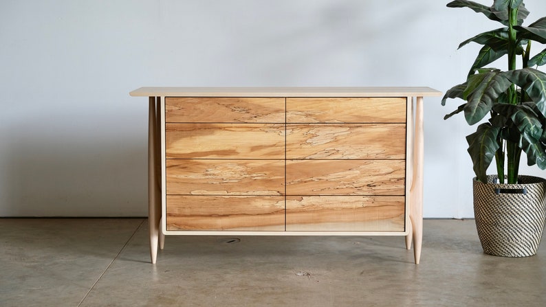 Op de afbeelding: Een lichte houten commode met zes laden en taps toelopende poten. De commode heeft een natuurlijke houten afwerking en een eenvoudig ontwerp.