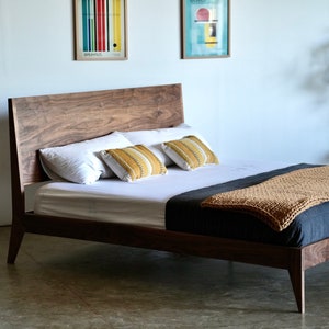 May include: A wooden bed frame with a dark walnut finish. The bed has a headboard with a slightly curved top and a low footboard. The bed is made with a simple, modern design. The bed is dressed with white bedding, two yellow and white striped throw pillows, and a tan knitted throw blanket.