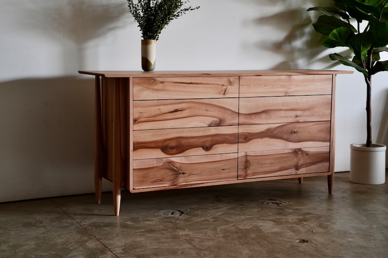 Może przedstawiać: Sześcio-szufladowa komoda wykonana z jasnego drewna z naturalnym usłojeniem. Komoda ma prosty, nowoczesny design z zwężającymi się nogami i lekko zaokrąglonym blatem.