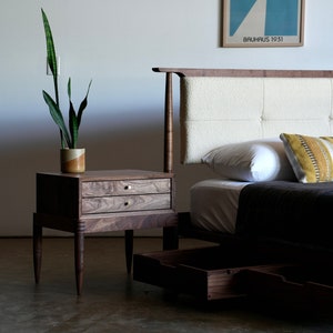 May include: A wooden bed frame with a white tufted headboard and two drawers. The bed has a dark gray blanket and two pillows with geometric patterns. A wooden nightstand with two drawers sits next to the bed, with a potted plant on top.
